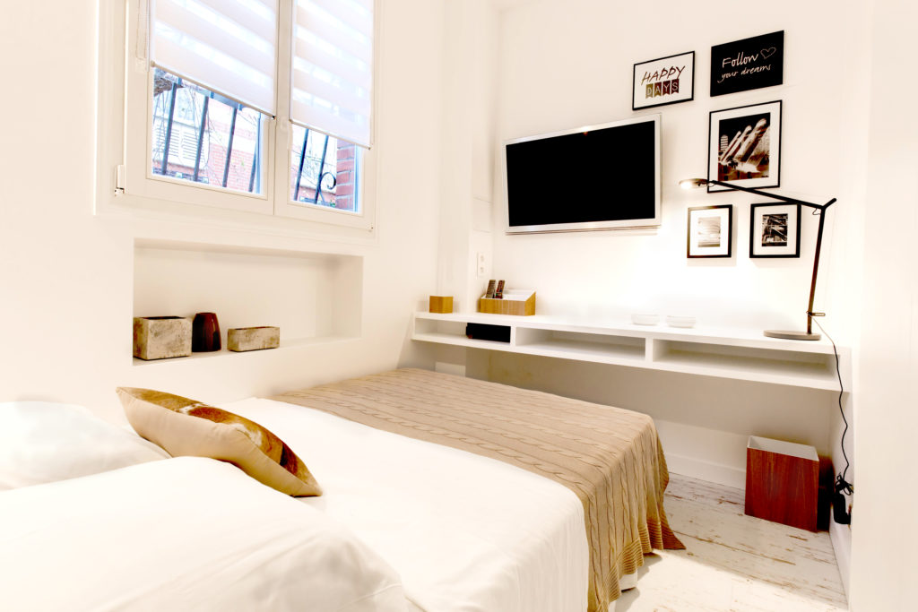 Une chambre avec un lit et sa couverte taupe face à une tv murale, des tableaux noirs et blancs au dessus d'une atagère blanche