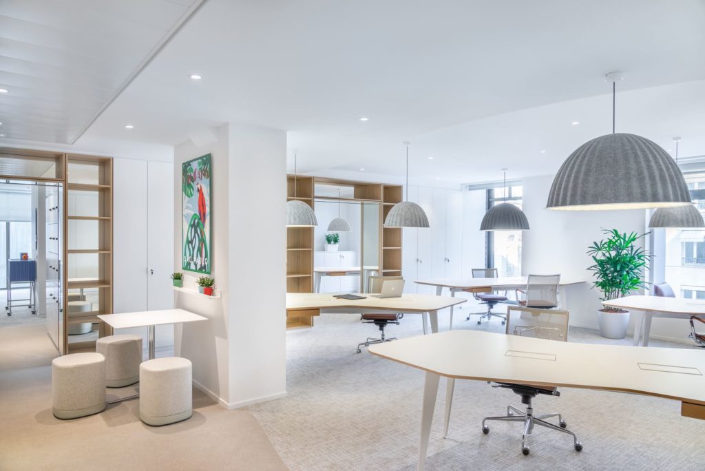 Un open space avec des tables de bureau ergonomiques des bibliothèques en bois et des sols en moquette. Le tout dans des tons clairs